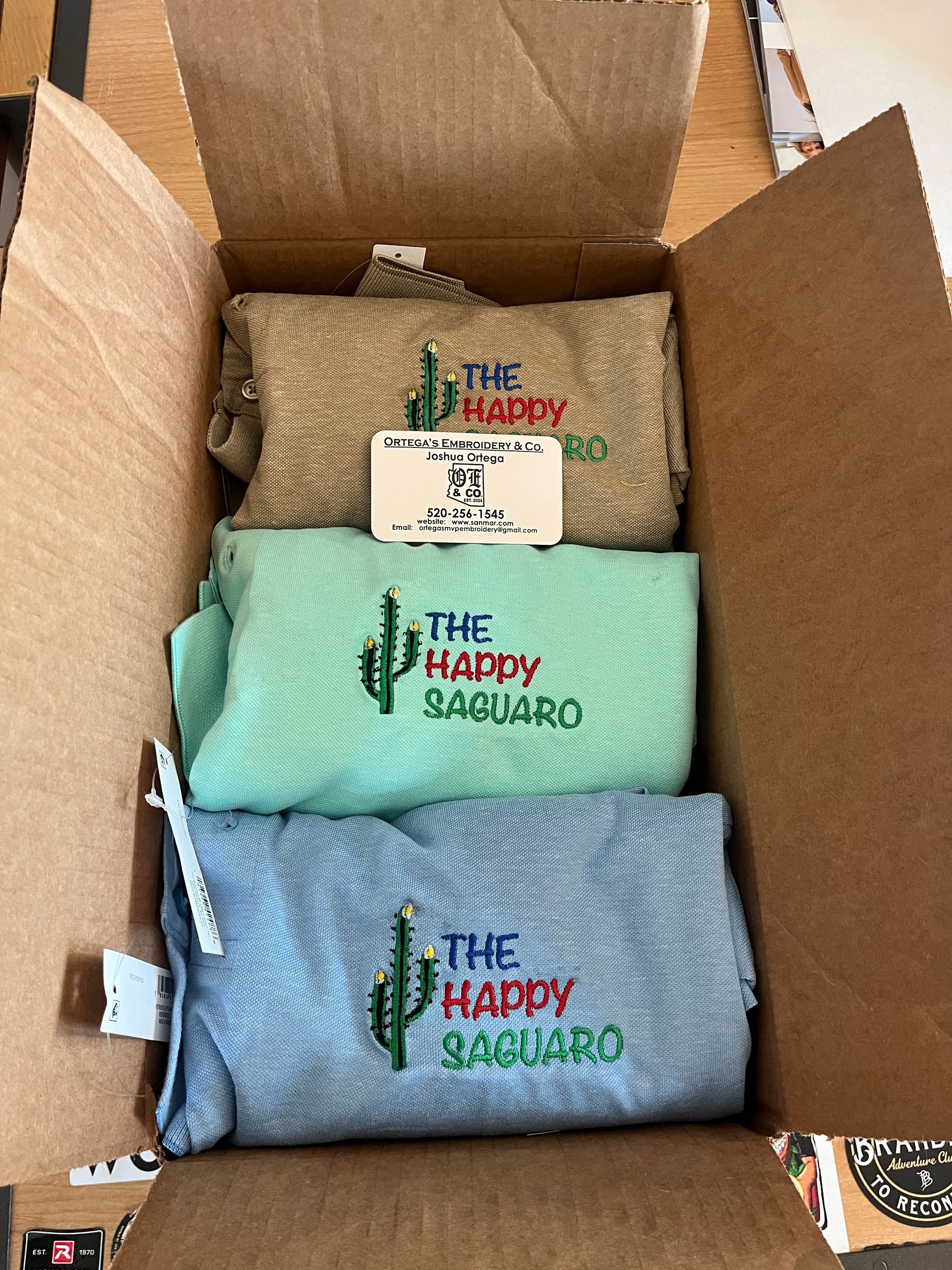 Three folded shirts in a box: tan, mint green, and blue, each with a cactus logo and text 