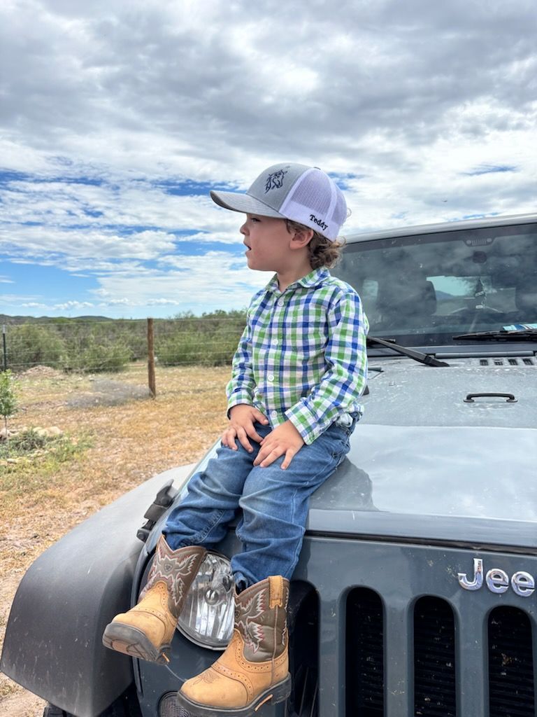 Child in cowboy boots and hat sits on a Jeep, looking toward the horizon.