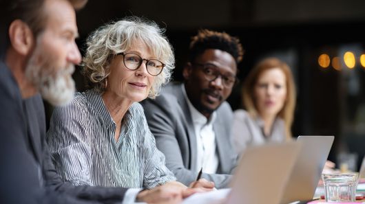 A group of professionals are sitting in a row with computers on the desk before them.