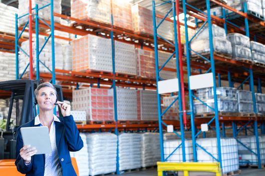 An executive stands in a warehouse with loaded shelves, talking on a headpiece