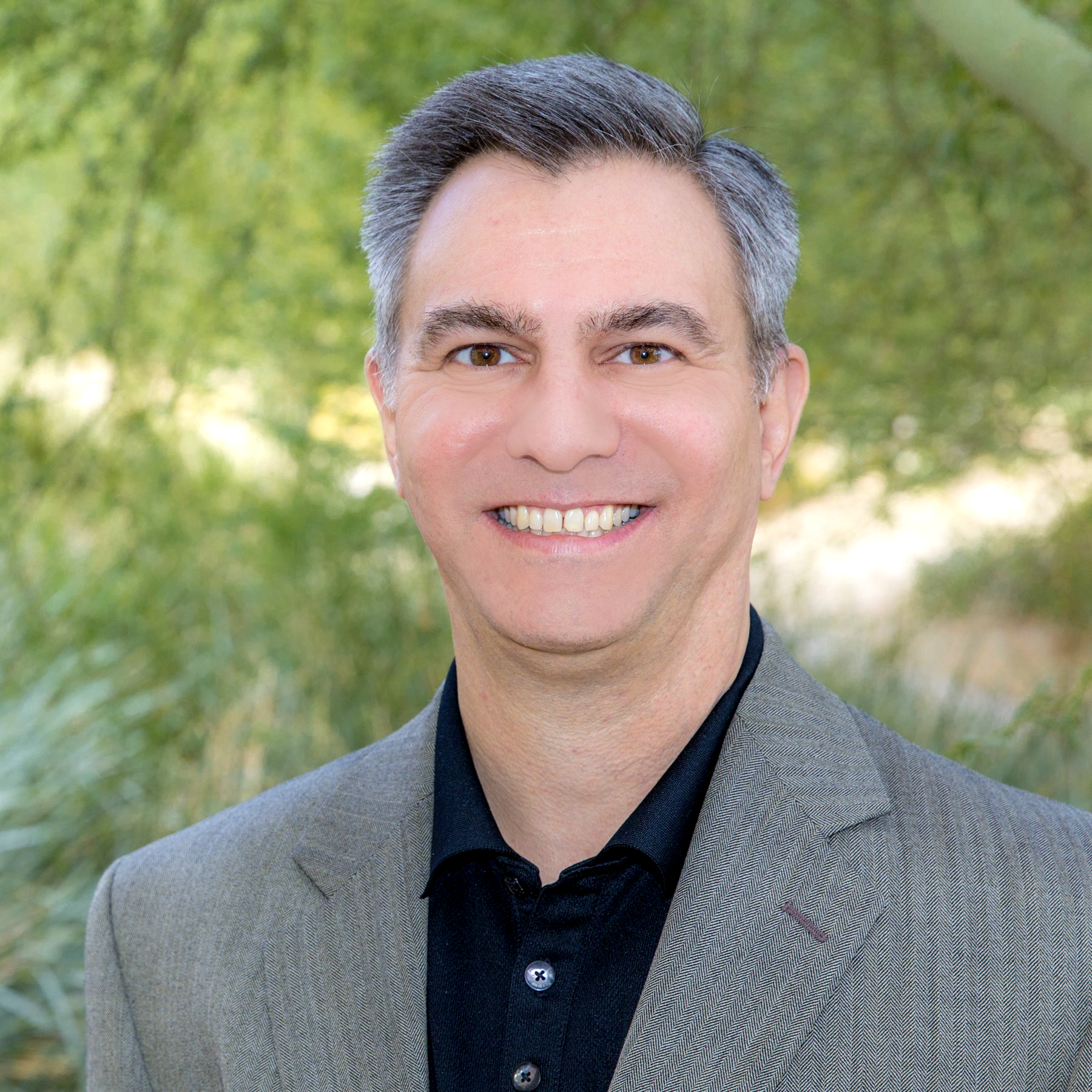 A grey haired man in a black and silver suit and black shirt is smiling for the camera.