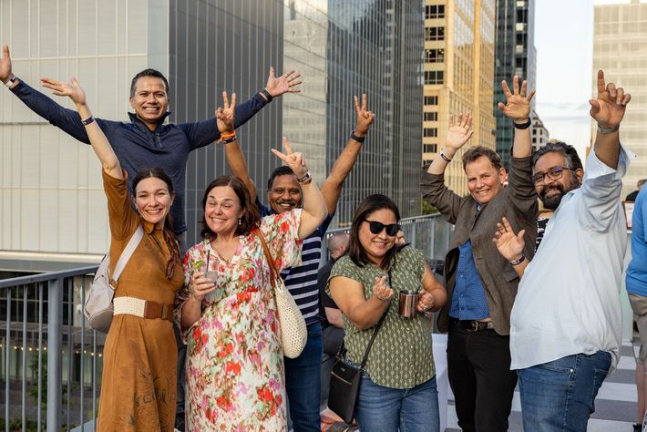 A diverse group of seven people stand on a rooftop bar in a city scape. They are posed with big smiles, giving peace signs and waving.
