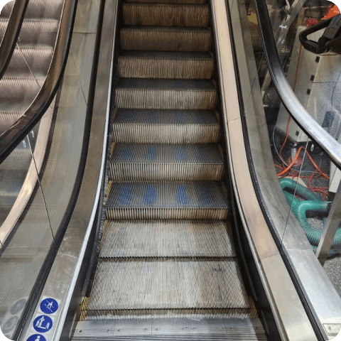 A close up of an escalator with a vacuum cleaner in the background.