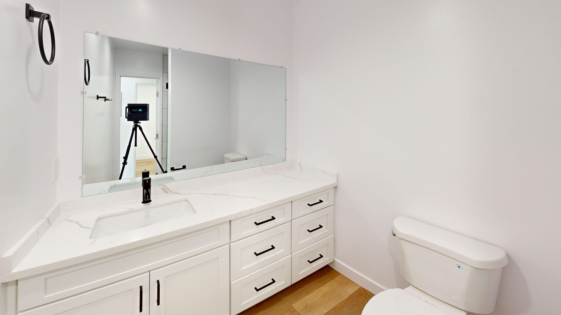 White bathroom with vanity, mirror, toilet, and towel rack. Camera on tripod in mirror's reflection.