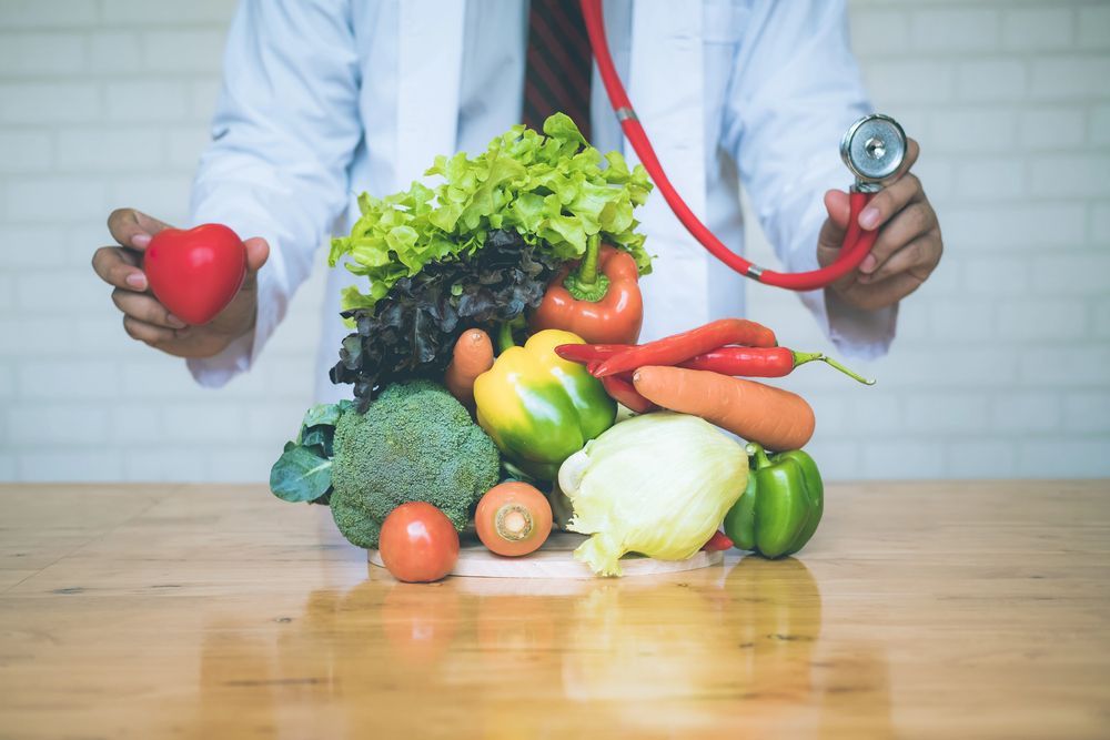 Un operatore sanitario tiene in mano un modello di cuore rosso e uno stetoscopio sopra un tavolo colmo di verdure fresche e colorate.