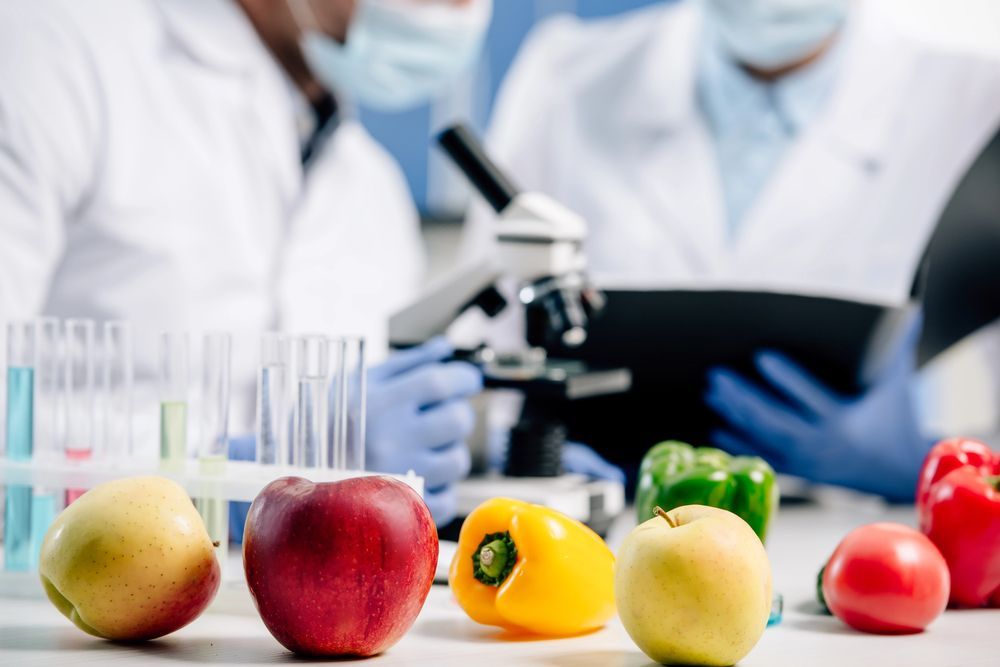 Due ricercatori in camice e guanti esaminano dei campioni in un laboratorio, con prodotti freschi sul tavolo in primo piano.