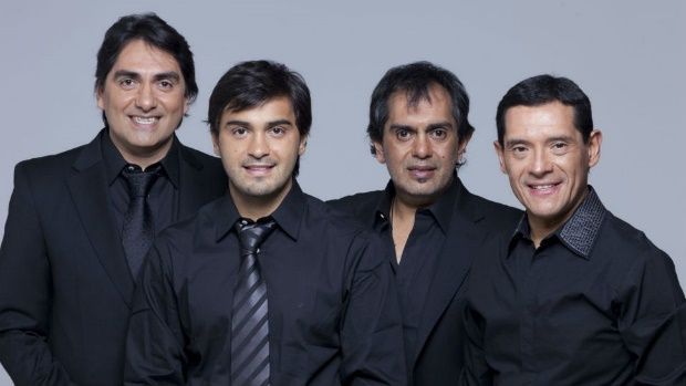 Cuatro hombres con camisas y chaquetas negras posando, sonriendo sobre un fondo gris.