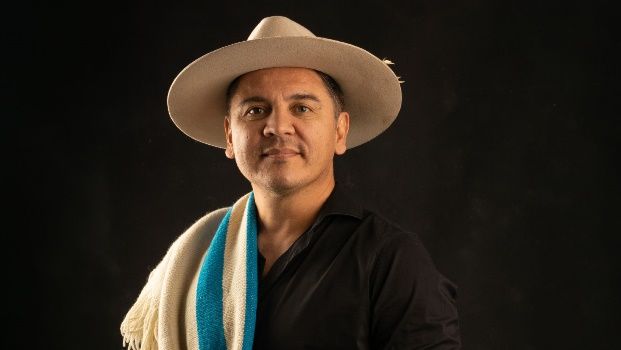 Hombre con sombrero y bufanda, sonriente, camisa oscura, sobre un fondo negro.