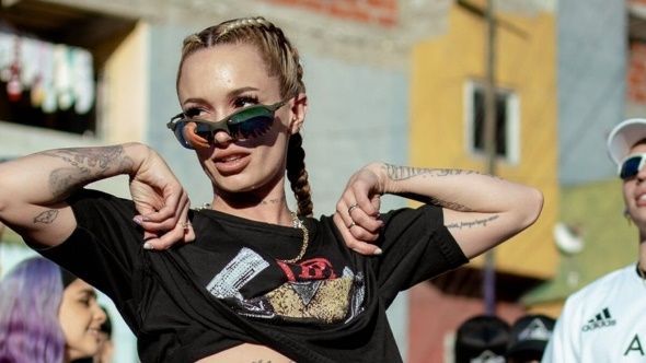Mujer con gafas de sol, trenzas y tatuajes apuntando hacia abajo frente a edificios coloridos.