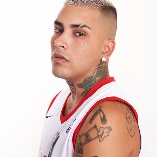 Hombre con tatuajes, piercings y cabello rubio, vestido con una camiseta blanca, mirando a la cámara.