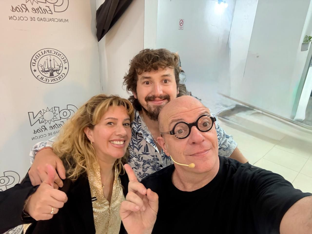 Tres personas sonriendo tomándose una selfi. Una señala hacia arriba, otra levanta el pulgar, con una pared blanca al fondo.