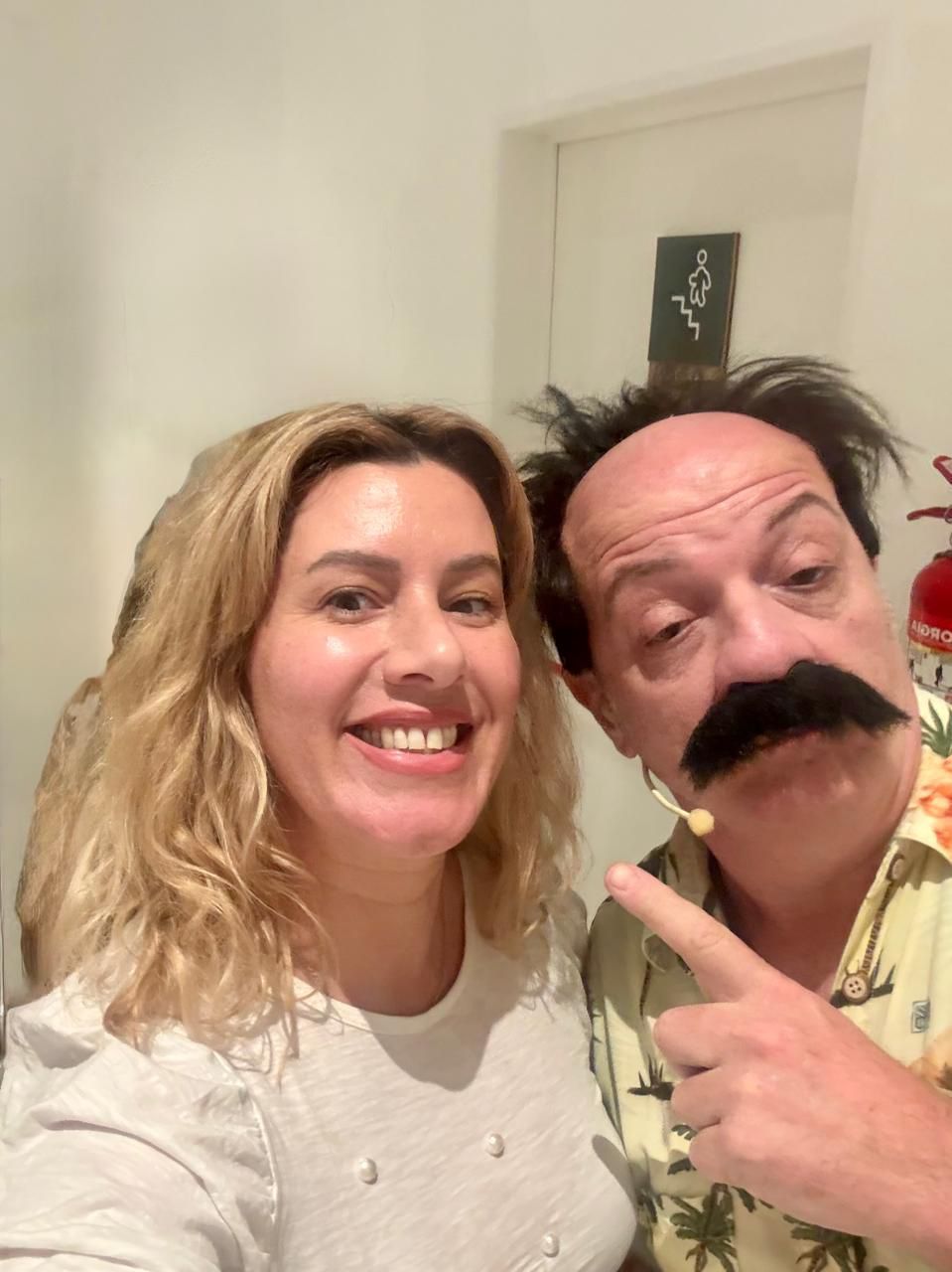 Una mujer rubia sonríe junto a un hombre con bigote postizo, señalándolo. Ambos en interiores.