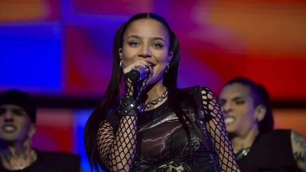 Cantante actuando en el escenario, vistiendo un traje negro con mallas, sosteniendo un micrófono, con bailarines de fondo.