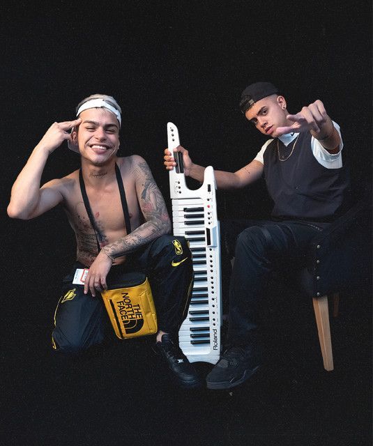 Dos personas posando: una con un keytar, sombrero y sonriendo; la otra señalando con la mano. Fondo negro.