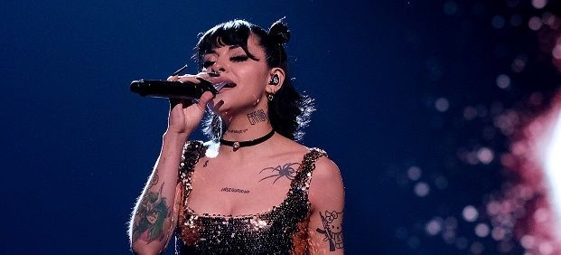 Cantante con tatuajes se presenta en el escenario, sosteniendo un micrófono, vistiendo un top brillante y una gargantilla.