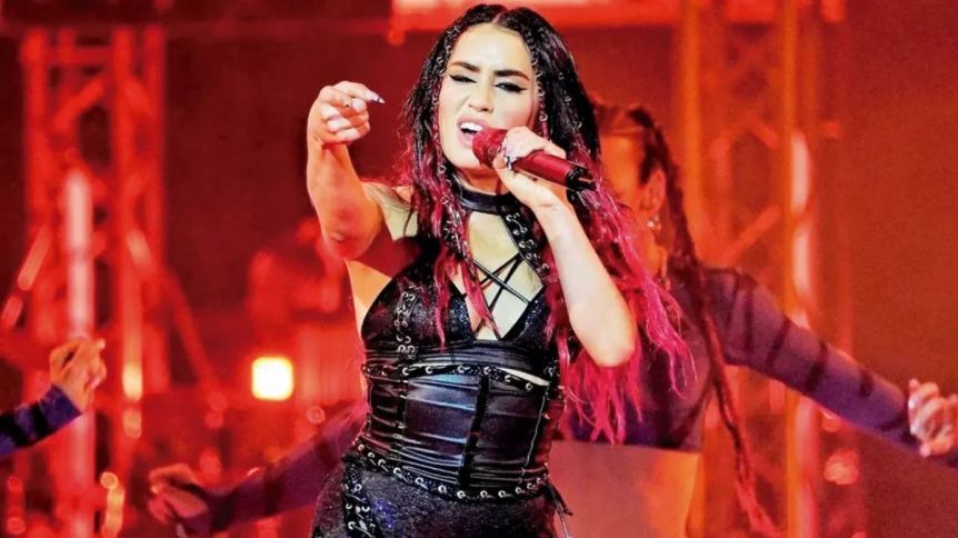 Cantante en el escenario, señalando, vistiendo traje negro con cabello rojo y micrófono, iluminación roja.