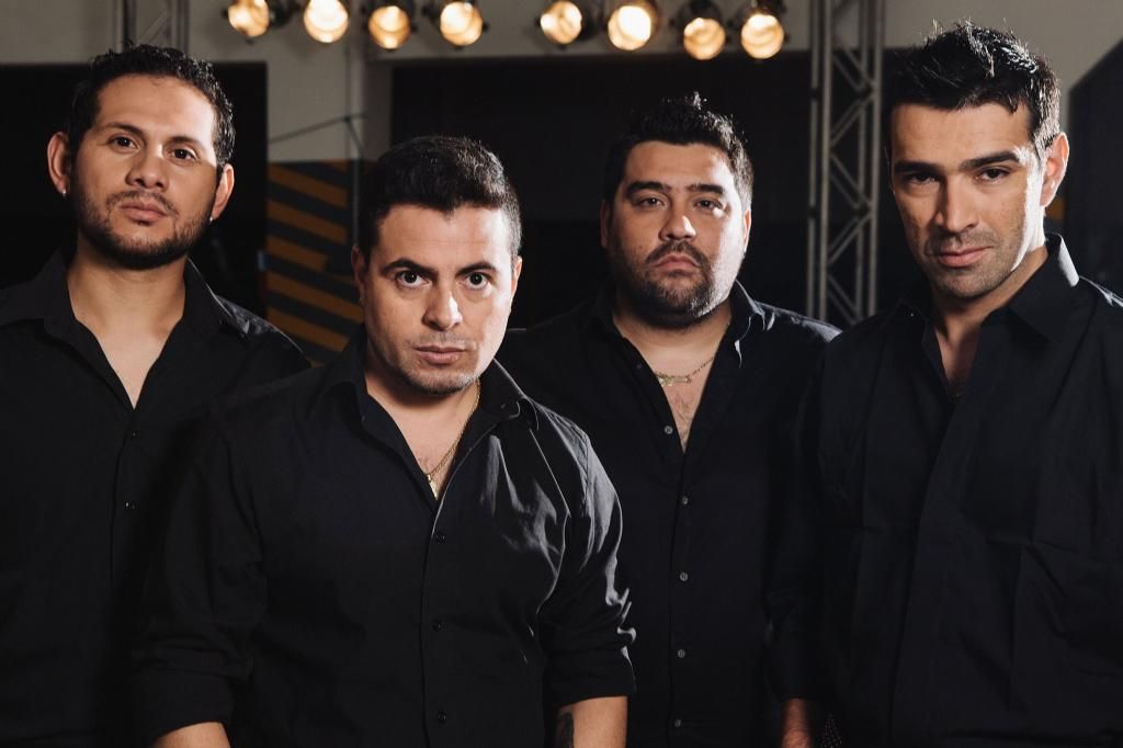 Cuatro hombres con camisas negras posan bajo las luces del escenario.