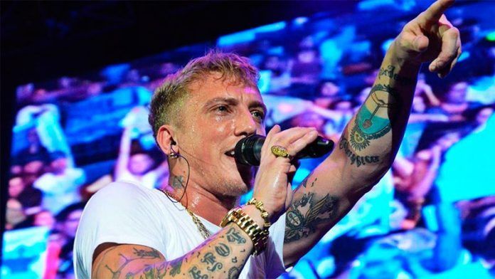 Hombre con tatuajes cantando en un micrófono en el escenario, con el brazo levantado.