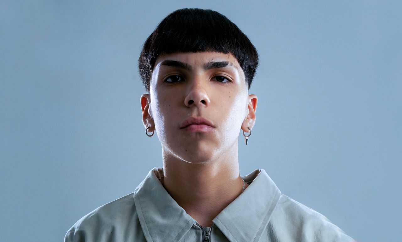 Persona joven con cabello oscuro, camisa de color claro y aretes, mirando hacia el frente.
