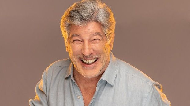 Hombre de cabello gris sonriendo ampliamente, vistiendo una camisa azul claro con botones.