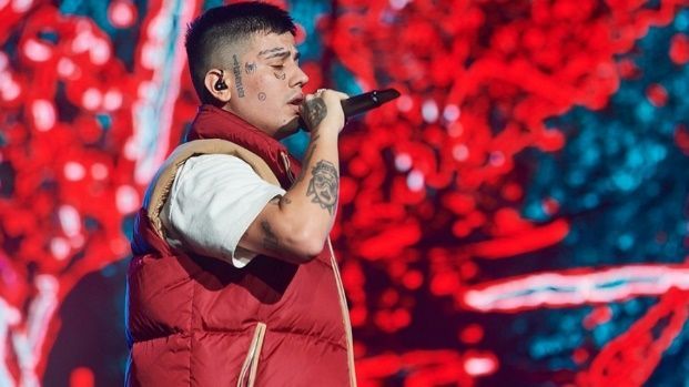 Hombre cantando en un micrófono en el escenario, vistiendo un chaleco rojo y una camisa blanca; fondo rojo y azul.