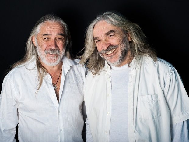 Dos hombres de pelo largo y gris sonríen, vistiendo camisas blancas sobre un fondo negro.