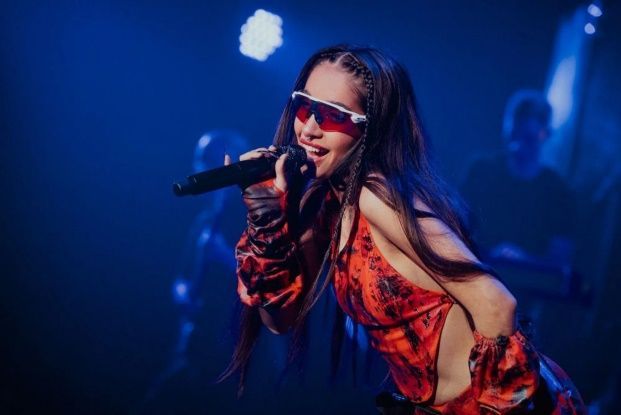Mujer cantando con un micrófono en el escenario, con gafas de sol y una camiseta naranja. Iluminación azul.