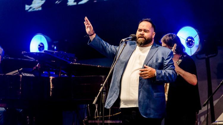 Hombre cantando con el brazo levantado, escenario iluminado con luces azules.