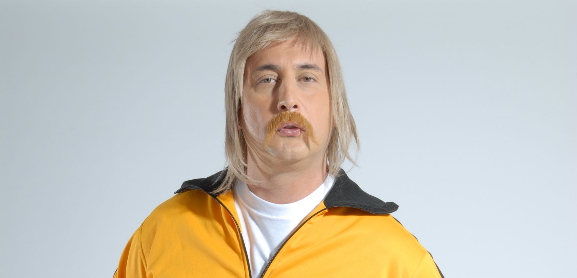Hombre con bigote, chaqueta amarilla y peluca rubia, sobre un fondo gris.