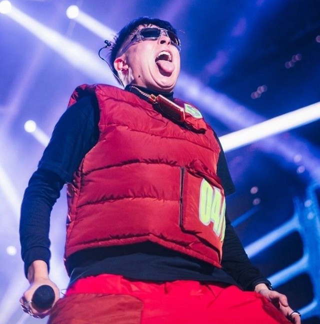 Persona en el escenario con gafas de sol, chaleco rojo y pantalones; boca abierta, lengua afuera, sosteniendo un micrófono.