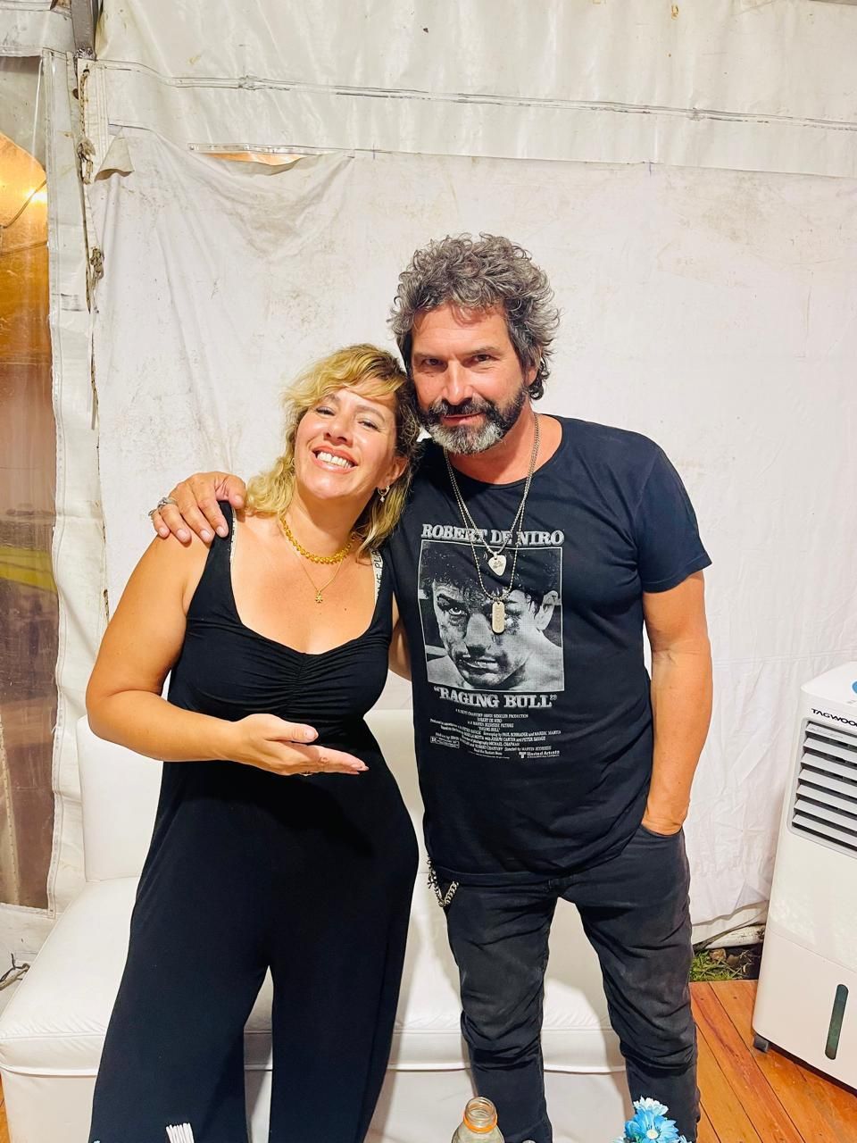 Una mujer con un mono negro y un hombre con una camiseta negra sonríen, tomados del brazo, dentro de una tienda de campaña.