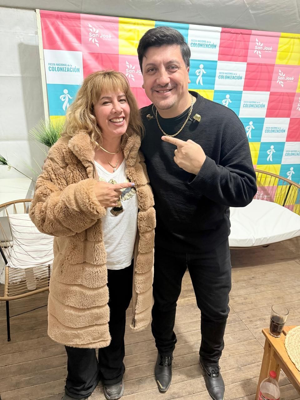 Dos personas sonriendo, posando para una foto: una mujer con abrigo y un hombre señalando. Fondo colorido.