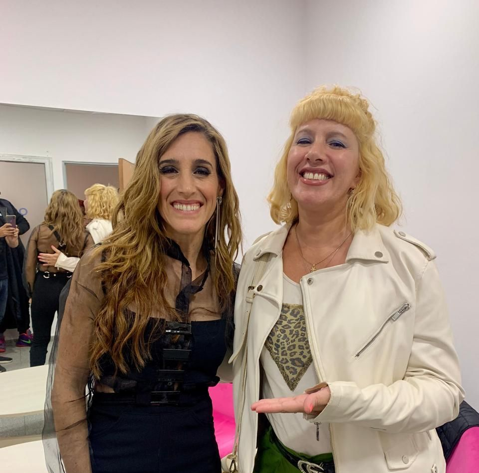 Dos mujeres sonrientes posan entre bastidores. Una lleva un traje negro y la otra una chaqueta blanca.