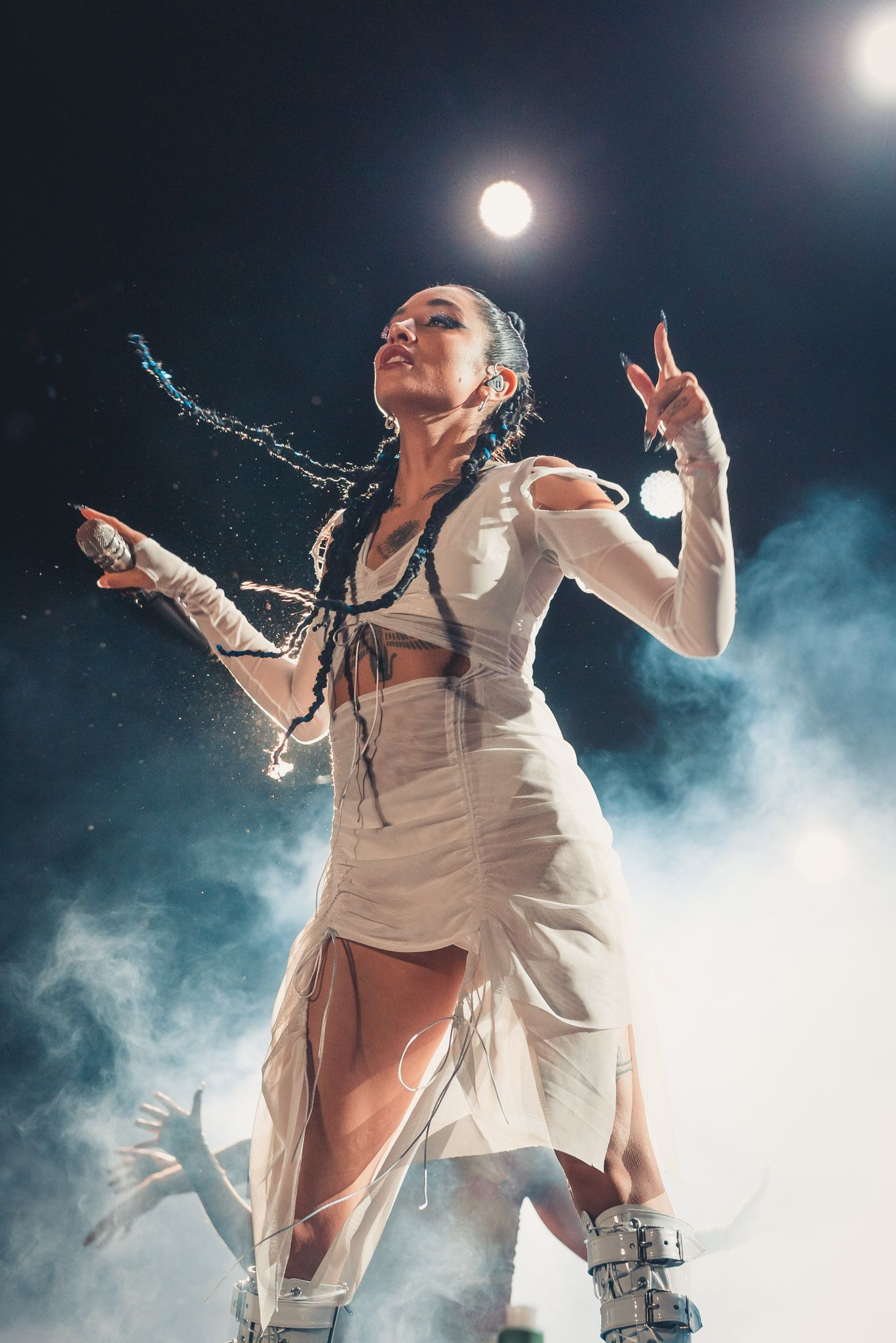 Mujer vestida de blanco actuando en el escenario, con el brazo levantado apuntando hacia arriba, humo.