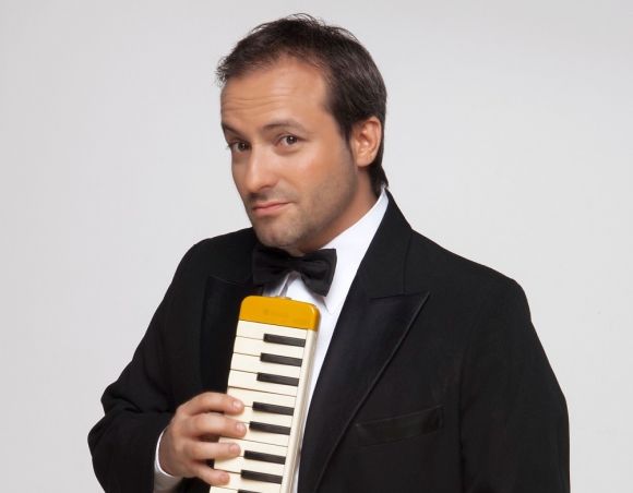 Un hombre con esmoquin sostiene un instrumento musical amarillo y blanco, sonriendo a la cámara.