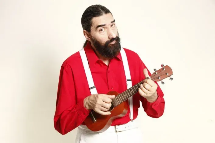 Hombre con camisa roja y tirantes tocando un ukelele, mirando al espectador.