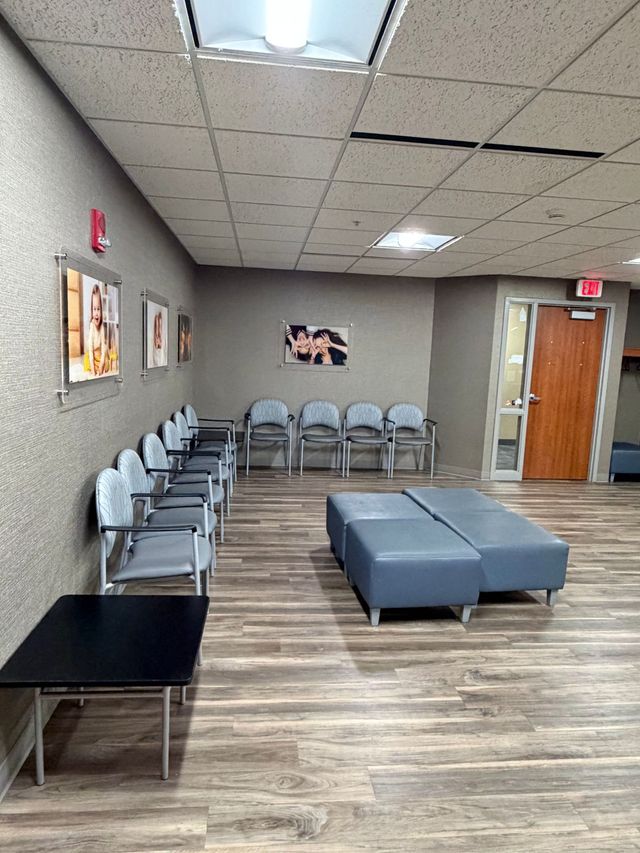 Waiting room with gray chairs, black table, blue ottoman, wood floor, and door with exit sign.