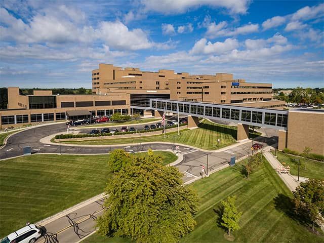 Large tan hospital building with curved entrance drive, green lawns, and blue sky with clouds