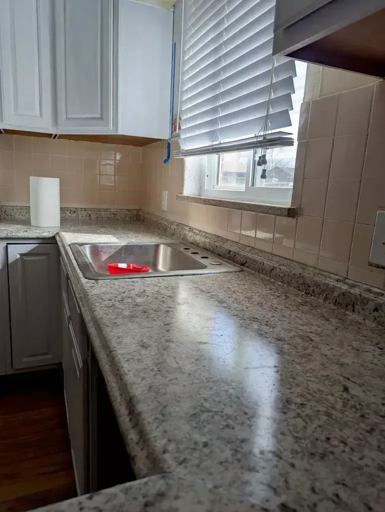 Kitchen countertop with sink and window; white cabinets and tan tile backsplash.