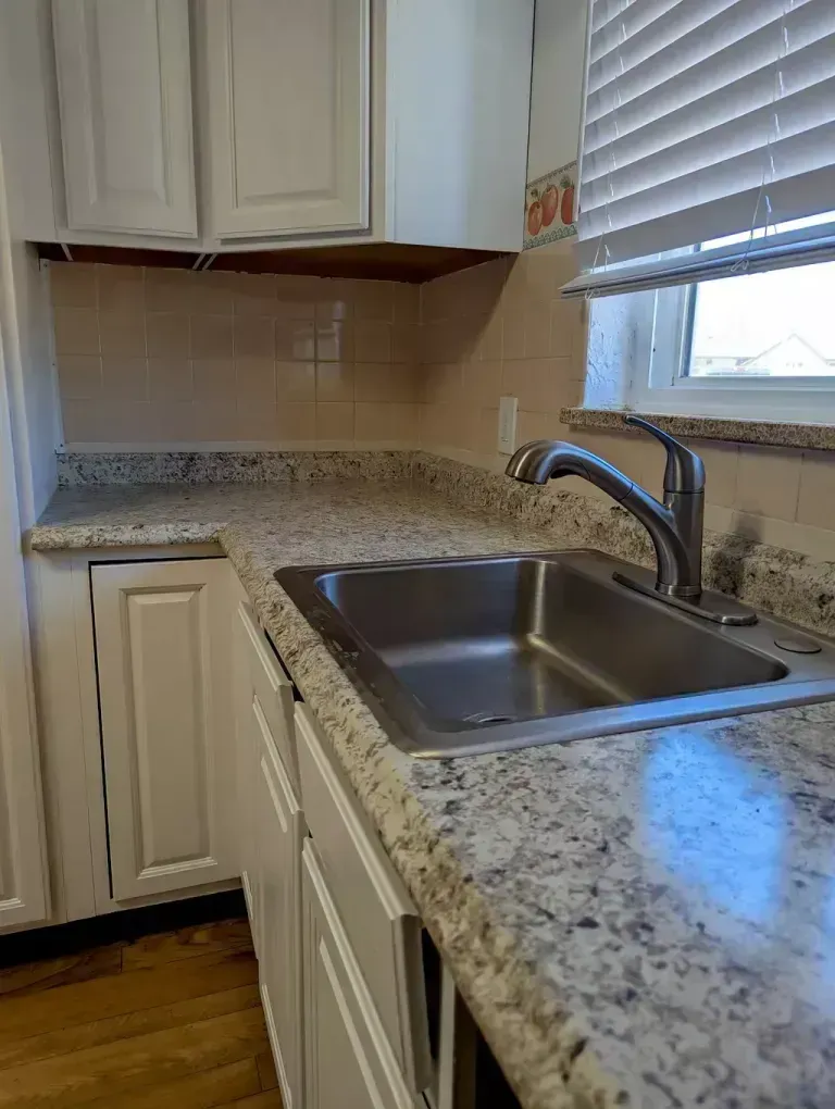 Kitchen with white cabinets, speckled countertops, stainless steel sink, faucet, and window.