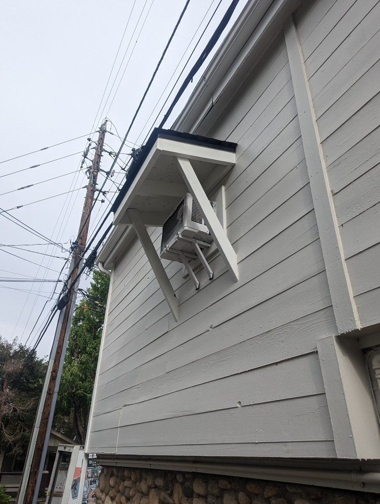 Exterior of a building with an air conditioning unit mounted on a bracket. Utility lines run overhead.