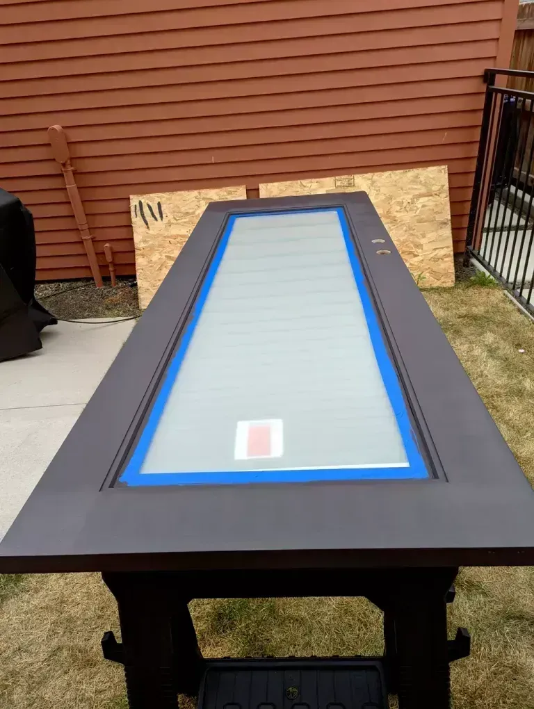Door with rectangular glass panel, painted dark brown, taped and resting on a sawhorse outdoors.