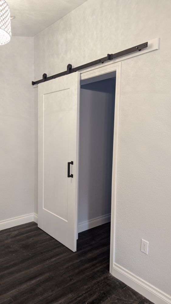 White barn door partially open, revealing a room with dark wood floor. Black hardware.