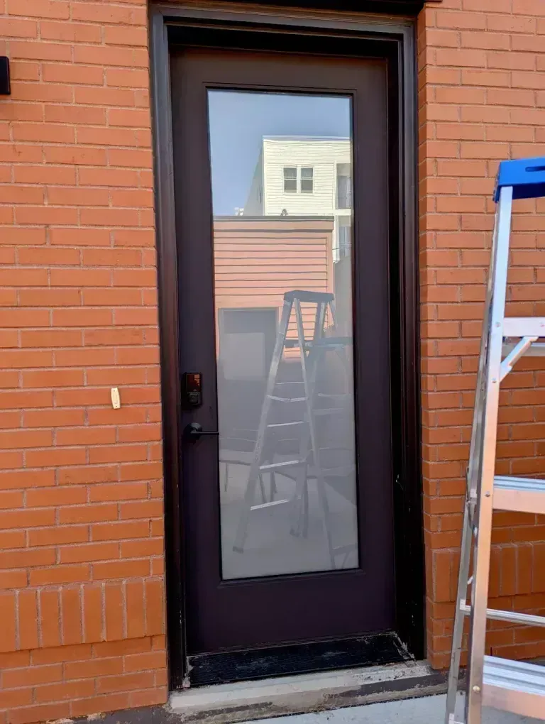 Black exterior door with a large glass panel set in a red brick wall, next to a silver ladder.