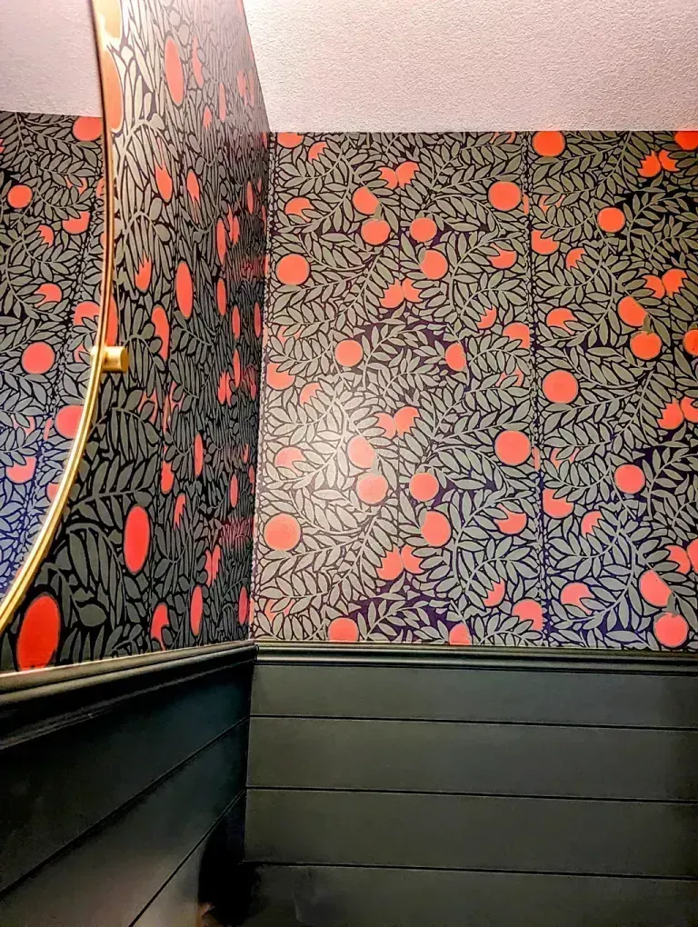 Corner of a room with floral wallpaper and dark green paneling. A gold-framed mirror is visible.