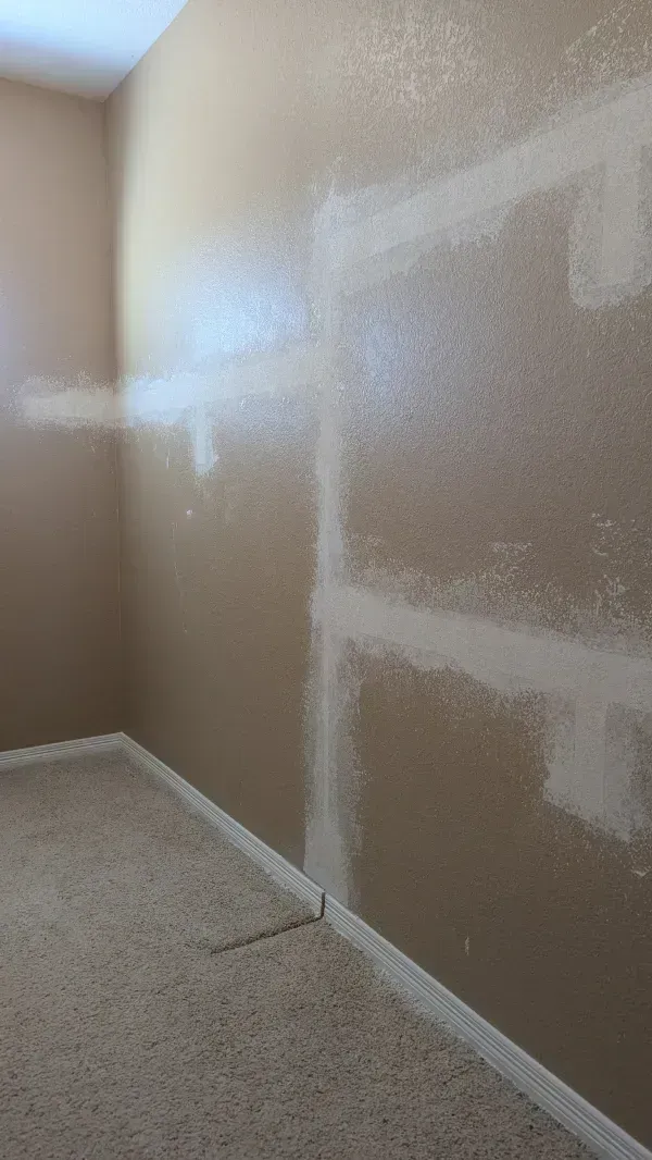 Interior view of a room with beige walls and carpet. White drywall compound is applied to the wall seams.