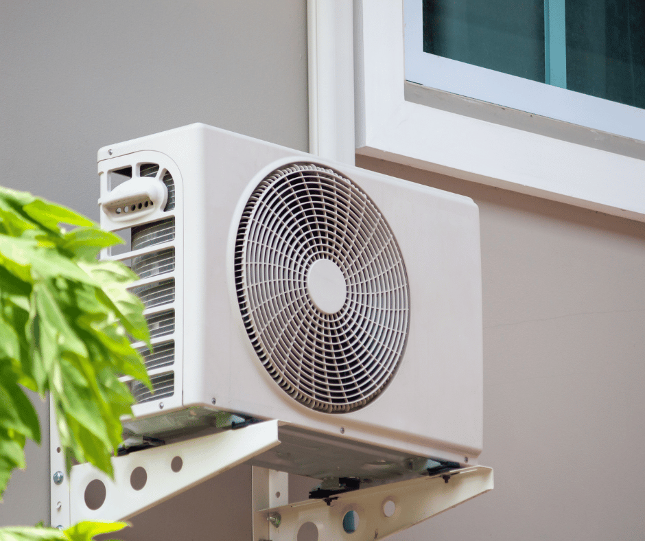A white air conditioner is mounted on the side of a building next to a window.