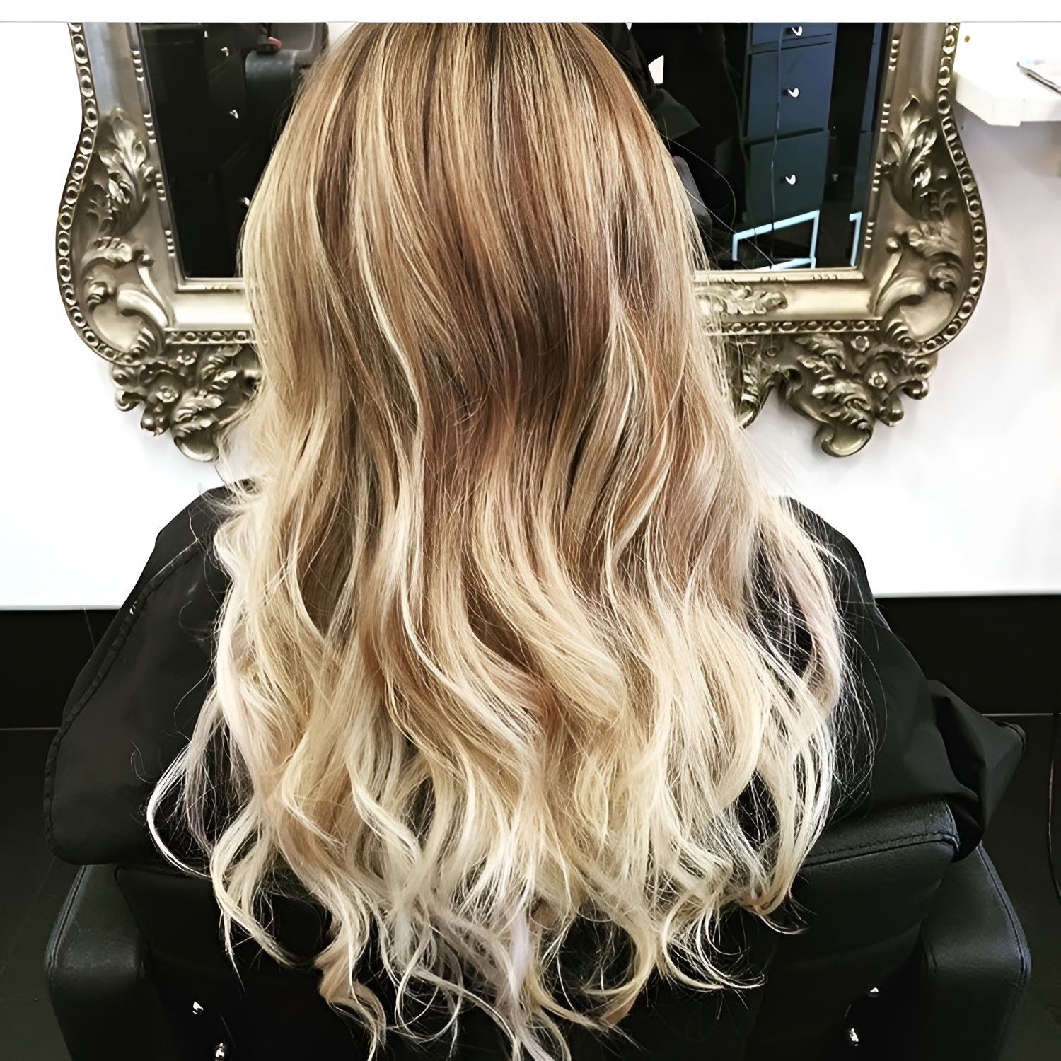 A person with wavy, shoulder-length hair styled in a blonde ombre, viewed from behind while seated in a salon chair.