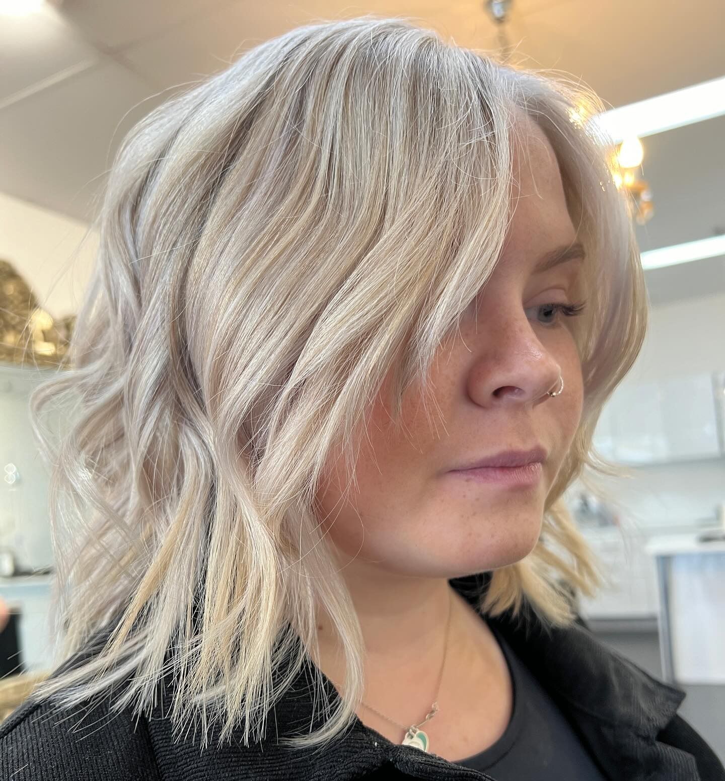 A person with shoulder-length, wavy platinum-blonde hair styled in a side part, set in a brightly lit salon.