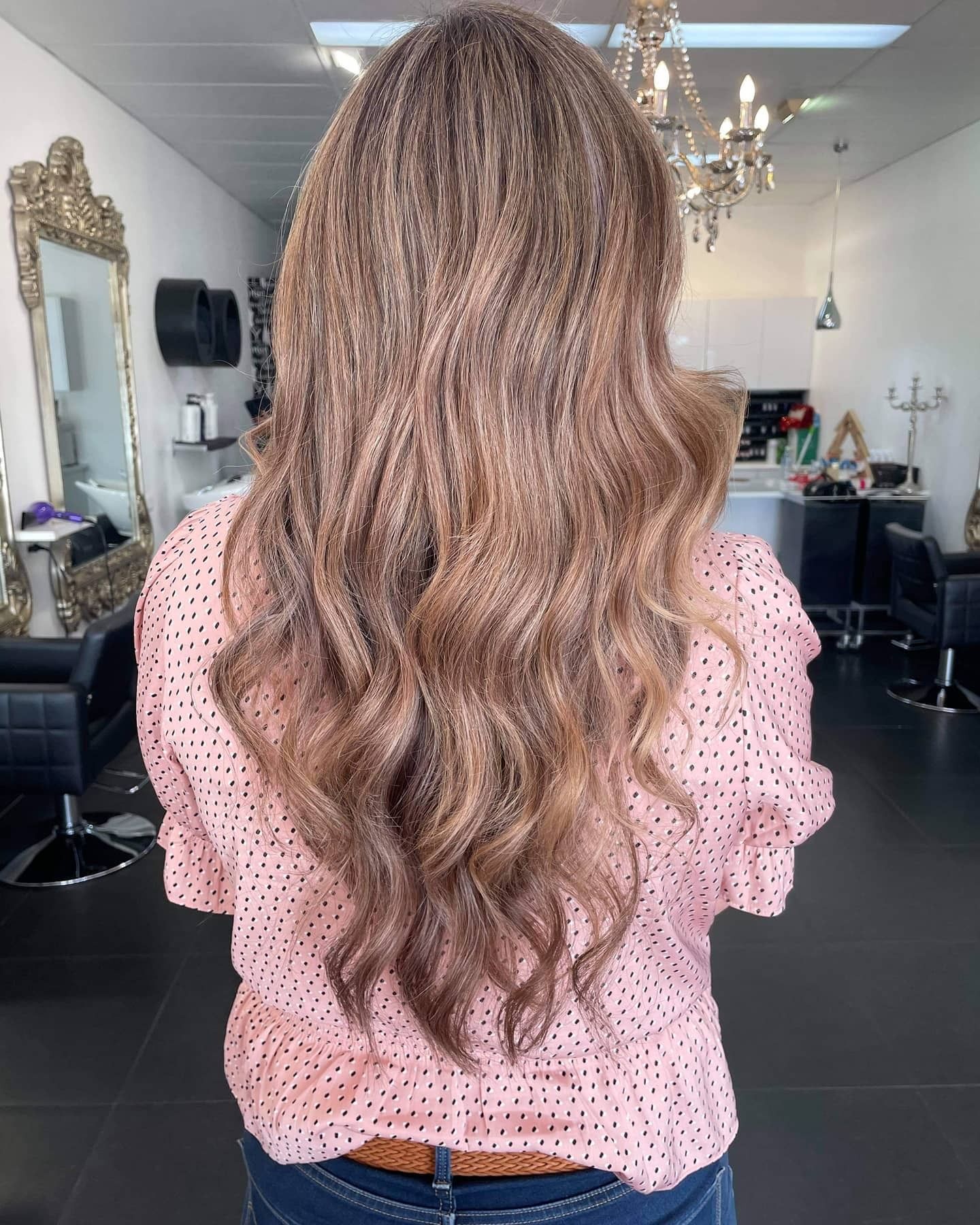 Woman with long wavy blonde hair standing in a salon, seen from behind, wearing a pink polka-dot blouse.